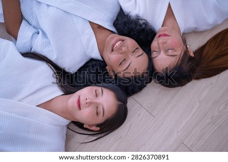 Top down shot of three young female friends resting side by side on a wooden floor in white spa robes as the woman with curly hair smiles faintly with her eyes closed.