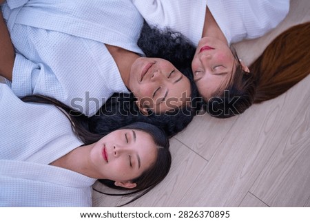Top down shot of three young female friends resting side by side on a wooden floor in white spa robes as the woman with curly hair lets her eyes fall shut in complete stillness.