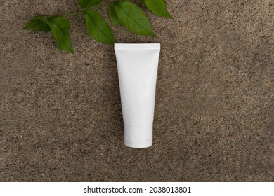 Top Down View Above Facial Skincare White Tube Bottle Product With Blank Label And Green Leaves On Sandy Dark Brown Stone Ground Rough Texture Background. Skincare Environmental Friendly Concept