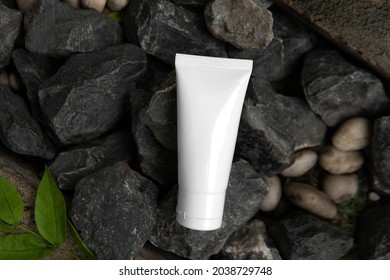 Top Down View Above Facial Skincare White Tube Product Blank Label On Crushed Dark Grey Stone Charcoal Color Or Road Metal Rough Texture And Green Leaf Background