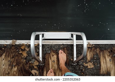 top down view of adult male feet at the edge of a heavily weathered dock with a shiny aluminium ladder leading into dark green murky lake water