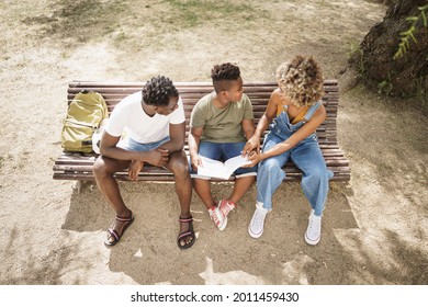 Top Down View Of African American Family Doing School Homework Sitting On A Bench In The Park. Little Son Doing School Work With Help Of His Parents