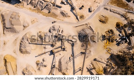 Top down view of aggregate processing machinery and roads. Conveyors, crushers and stockpiles form an industrial layout with loader tracks, dust and surrounding trees.
