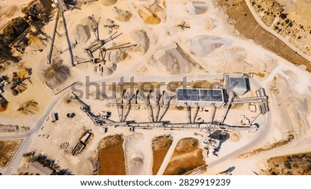 Top down view of aggregate processing plant and stockpiles. Conveyor lines connect sorting units, storage bays and solar panel roofs among pale sand piles and service roads.