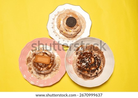 Top down view of an assortment of three gourmet cinnamon buns on colorful plates with different sweet toppings on a solid bright yellow background