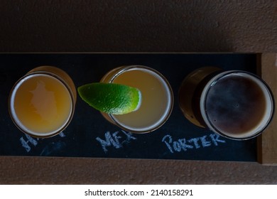 A Top Down View Of A Beer Flight Of A Variety Of Craft Beers. The Small Clear Glasses Range In Golden Colors And Are Samples Of IPA, Mexican, And Porter.The Glasses Are On A Dark Wood Board.