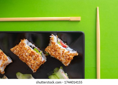 Top Down View Of A Black Square Plate With Multiple Sushi Rolls With Sesame Seeds And Cucumber On Green Simple Background With Chopsticks Paralel To The Plate