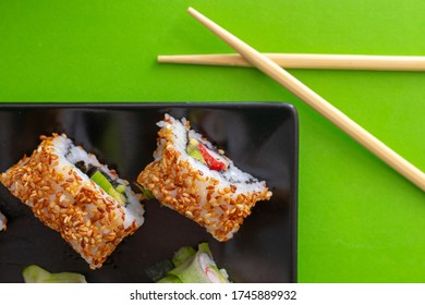 Top Down View Of A Black Square Plate With Multiple Sushi Rolls With Sesame Seeds And Cucumber On Green Simple Background With Crossed Chopsticks