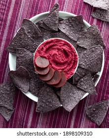 Top Down View Of A Bowl Of Beetroot Hummus Surrounded By Blue Corn Chips.