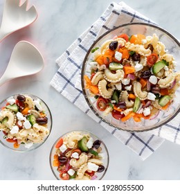 Top Down View Of A Bowl Of Greek Pasta Salad With Two Smaller Serving Bowls Of The Salad.