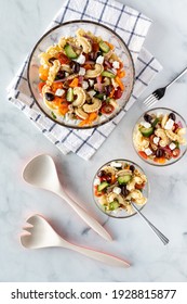 Top Down View Of A Bowl Of Greek Pasta Salad With Two Servings Of The Salad And Salad Serving Utensils.