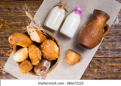 Top Down View Of Bread Basket Full Of Loaves Next To Milk Bottles, Pitcher And Cup On White Paper Over Old Wooden Table