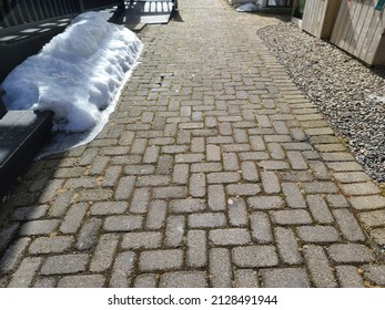 A Top Down View Of A Brick Walk Way. There Are Piles Of Snow Melting To The Side Of The Pavers. There Is Moss Growing In Between Each Brick.