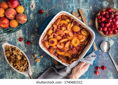 A Top Down View Of A Casserole Dish Of Delicious Baked Stone Fruit Cobbler With A Hand And Napkin Holding One End.