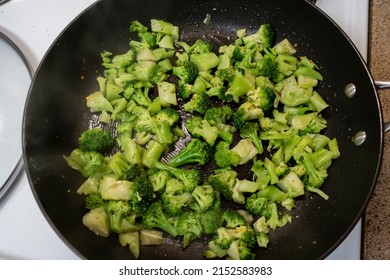 Top Down View Of Chopped Up, Frozen Broccoli Cooking In A Large Black Skillet On A White Stove Top