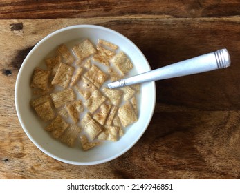 Top down view of cinnamon cereal squares in a bowl with milk. White bowl against a wooden table with a metal spoon in the cereal bowl. 