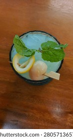Top down view of a cold blue cocktail garnished with fresh mint leaves, lemon slice, and a lychee on a wooden table.