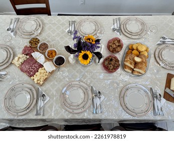 The top down view of a dining room table set for the holidays. There is fine china, a meat and cheese platter, corn muffins, Italian bread, and crackers.
