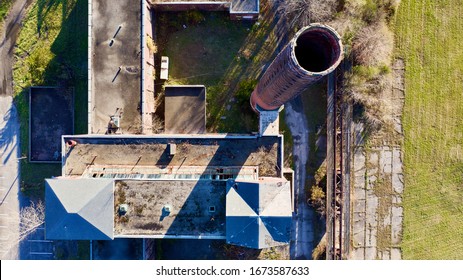 A top down view directly above an abandoned building with a tall, inactive smoke stack. The drone camera is high up and looking down into the smokestack. It was taken on a sunny day.