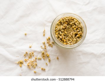 Top Down View Of Dried Chamomile Flowers For Tea In Glass Jar And Scattered On White Tablecloth (selective Focus)