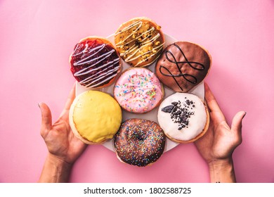 Top Down View Of Female Hands Holding Dessert Plate Full Of Colorful Donuts, Isolated On Top Of Pink Background. 