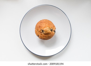 Top Down View Of A Fresh Uneaten Blueberry Muffin On A Plate Against A White Background With Natural Morning Light.