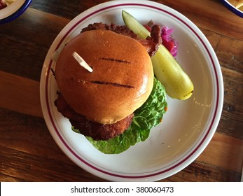 Top Down View Of A Fried Chicken Burger In A Semi Charred Brioche Bun With Pickle On A White Plate On A Wooden Table In A Restaurant