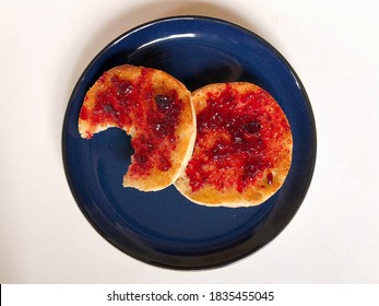 Top Down View Of Fruit Jam On English Muffin On A Plate. A Bite Is Eaten From One Piece Of Bread On A Table.