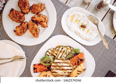 Top Down View Of Greek Food: Tomato Balls ( Tomato Fritters, Tomatokeftedes), Tzatziki Sauce With Olive Oil And Grilled Cheese With Vegetables In Greece, Santorini Tavern Table.