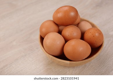 A top down view of a group of fresh organic brown chicken eggs arranged inside a rustic wooden bowl on a textured light wood surface