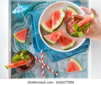 Top Down View Of A Hand Holding A Watermelon Mocktail With A Tray Of Watermelon.