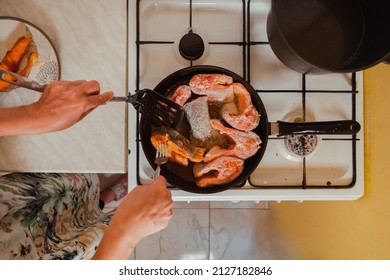 Top Down View Hands With A Fork And A Skimmer Frying Salmon Fish In A Hot Pan On A Gas Stove At Home