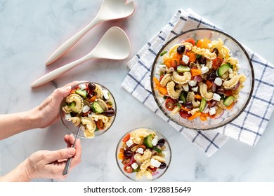 Top Down View Of Hands Holding And Helping Themselves To A Bowl Of Greek Pasta Salad.