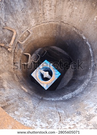 Top down view looking into a deep concrete utility manhole shaft with rusty metal steps and a blue fire hydrant at the bottom.