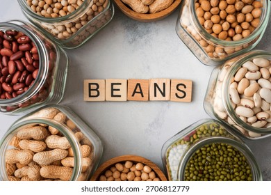 A top down view of multiple glass jars filled with different types of beans and legumes, arranged in a circle around the word BEANS.