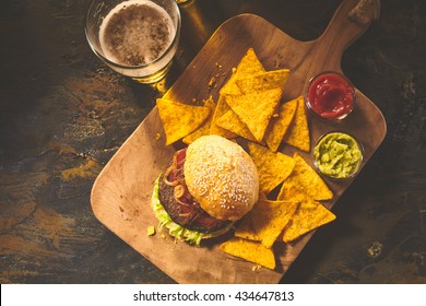 Top Down View On Burger In Sesame Bun, Tortilla Chips, Salsa And Guacamole On Cutting Board With Beer On Table