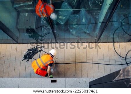 Top down view on a construction site worker in orange high visibility vest and white hard safety hat cutting electric cables to a specific size. Selective focus. Wiring a house concept.
