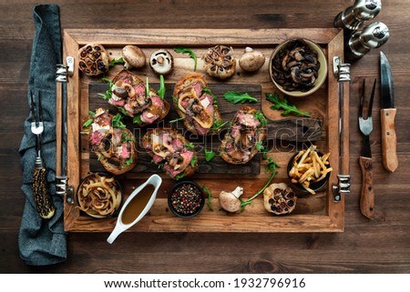 Top down view of open faced steak sandwiches on a wooden platter surrounded by toppings.