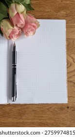 Top Down View Of Open Notebook With  Pen And Pink Roses On Wooden Desk