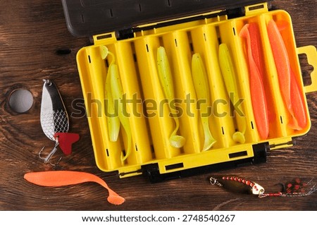 Top down view of an open yellow tackle box filled with colorful soft lures, with metal spoons placed on wooden surface nearby