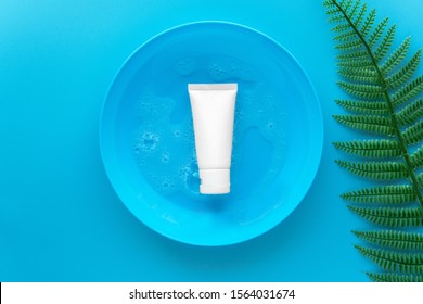 Top Down View Of Organic Gentle Skincare Product Hyaluronic Acid Facial Cleanser White Tube For Dry Skin On A Blue Dish With Soap Water Decoration With Artificial Fern Leaf On Plain Blue Background.