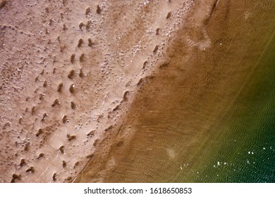 A Top Down View Over A Beach, Directly Above The Shoreline. You Can See Foot Prints In The Sand, Creating An Interesting Pattern. You Also See Shades Of Green In The Waters Approaching The Shore.