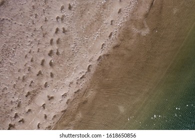 A Top Down View Over A Beach, Directly Above The Shoreline. You Can See Foot Prints In The Sand, Creating An Interesting Pattern. You Also See Shades Of Green In The Waters Approaching The Shore.