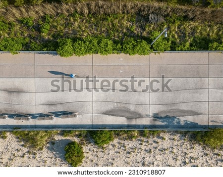 A top down view over the concrete boardwalk at Rockaway Beach in Queens, NY on a sunny day. The camera is tilted straight down as people enjoy the day, casting their shadows onto the ground.