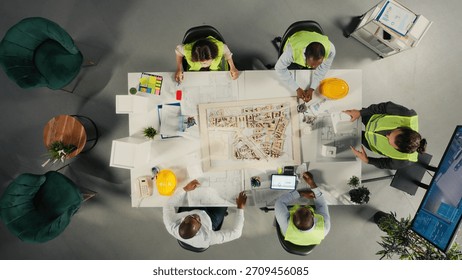 Top down view of property developers and architects in meeting reviewing blueprints and structural designs. Team discusses project approval and city planning for housing growth.