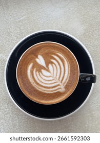 Top down view of single cup of coffee with leaf latte art inside a black ceramic cup and white marble table in the background