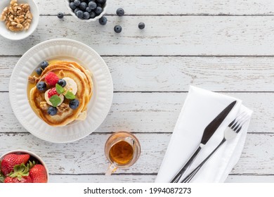 Top Down View Of A Stack Of Buttermilk Pancakes Topped With Fruit, On A Wooden Table And Cutlery To The Side.