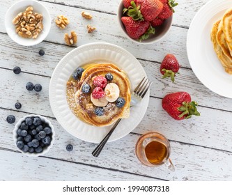 Top Down View Of A Stack Of Buttermilk Pancakes Topped With Nuts, Fruit And Maple Syrup And Sprinkled With Powder Sugar.