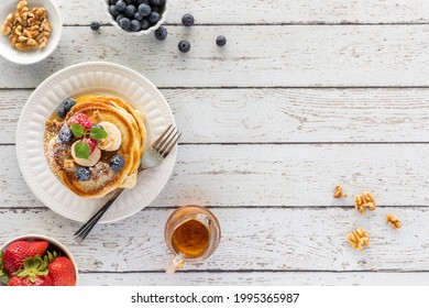 Top Down View Of A Stack Of Buttermilk Pancakes Topped With Berries, Banana Slices, Syrup And Powdered Sugar.