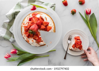 Top Down View Of A Strawberry Shortcake With A Hand Holding A Plate With A Slice Of Cake On It.